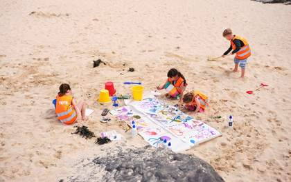 Children in Tate orange hi-vis vests on Porthmeor beach. One child is making patterns in the sand with a broom. Two children are painting sea creatures with inks on a large sheet of paper on the sand.