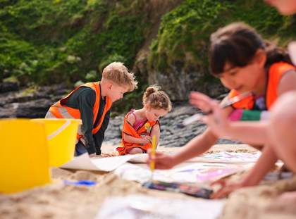Children in Tate orange hi-vis vests on Porthmeor beach. They are all painting on a large sheet of paper on the sand.