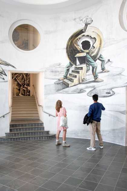 A couple of people seen from behind in a round room with a staircase looking at a surreal artwork on the wall.