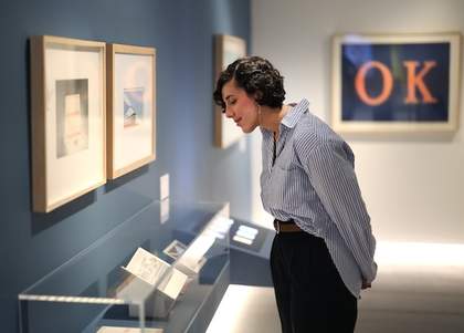 A photograph of a person looking down at some artwork on display in a glass cabinet. There is artwork on the wall in the background.