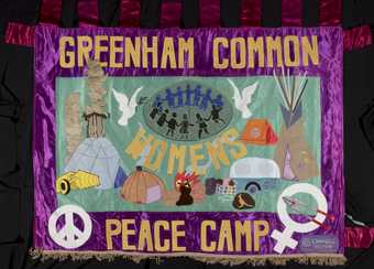 A quilt, made up of different colours including purple, blue, yellow and white, with a dove, a tent a fire, a caravan and a peace symbol embroidered on the material.