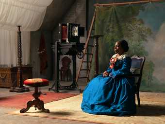 A still from 'lessons of the hour' by Isaac Julian. A woman of colour sits in a chair in a blue dress against a false backdrop of tress and blue sky. She stares at a footrest opposite her.