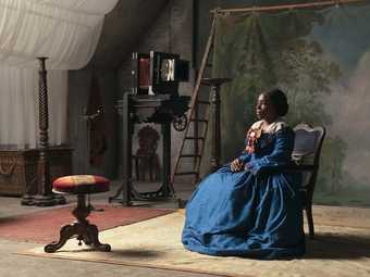 A still from 'lessons of the hour' by Isaac Julian. A woman of colour sits in a chair in a blue dress against a fake backdrop of tress and blue sky. She stares at a footrest opposite her.