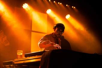 A photograph of a person at a DJ deck in a dark room with bright orange/yellow lights.