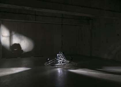 Dimly lit industrial room with concrete walls and a stack of metal that looks like rigging in the centre of the floor.