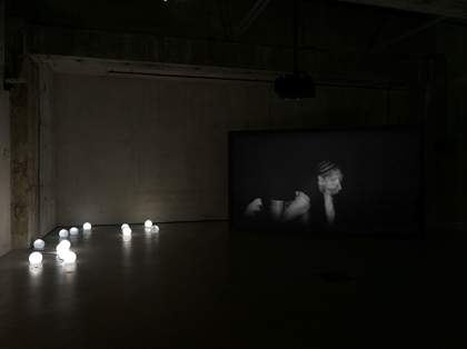 Dimly lit room, industrial concrete walls with a few small globe shaped lights on the floor. A blurred image of a woman, head leaning on her hand is on a screen in the corner.