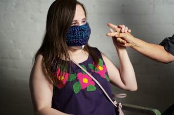 A photograph of Theresa Lambert wearing a purple tank top with pink flowers and a face mask woven with purple wool. Her hand is pictured touching another persons hand using American Sign Language..