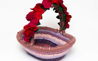 A hand woven basket in different shades of pink and purple, the handle is covered in bright red felt flowers. This is an artwork by Theresa Lambert who is speaking at the upcoming event with Abbas Zahedi.