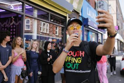 A photograph of a young man holding his phone out as he records himself standing in front of a group of young people.