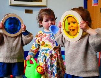 Three children, the one on the left looking through a blue colour lens, one in middle holding a watering can and a toy boat, the one on the right looking through a yellow colour lens