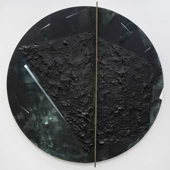 A dark circular abstract sculpture mounted on a white wall. A collage of textures in black and very dark green with a thin brass piece crossing the circle off-centre.