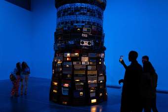 Silhouetted people in a dark blue room looking at an artwork that is a tower made from radios