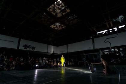 A performer dressed in yellow stands in the middle of a darkly lit stage, surrounded by an audience.