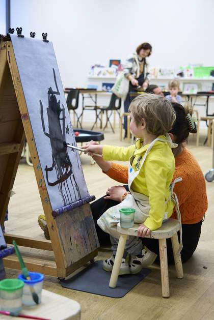 A young boy paints on paper with black ink, sitting at an easel, being helped by a woman