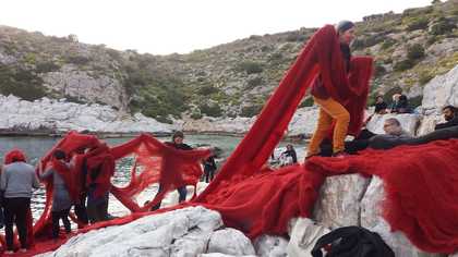 A group of people are moving a large piece of red fabric across white rocks in a beach landscape.