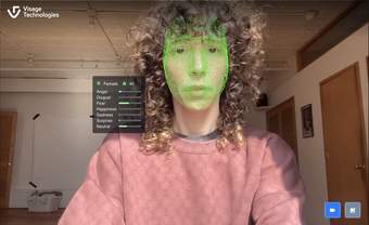 A female face looking a camera within her home with a bright green mesh mapped to her face and a pop-up window measuring her emotions to the left.
