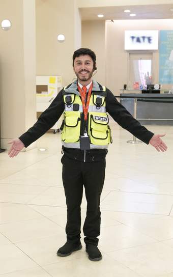 A person wearing a high visibility jacket, with arms open in a welcoming pose and smiling