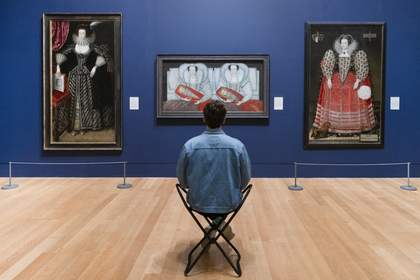 A person sitting on a foldable stool in an art gallery with dark blue walls