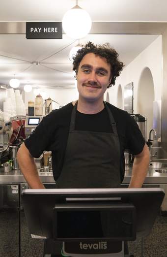 A person with dark curly hair and a moustache wearing a black t-shirt and a black apron