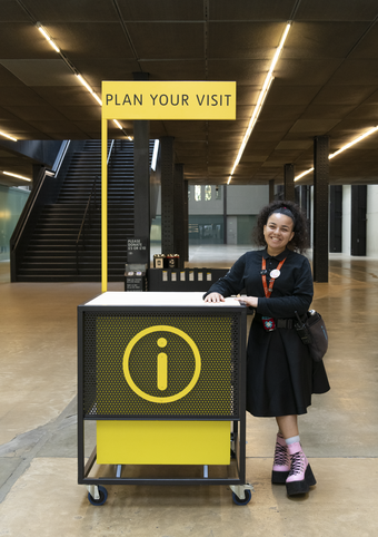 A person standing and smiling leaning on a desk that says Plan Your Visit