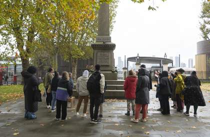 A large group of people stand outside, surrounding a large memorial statue, listening to a tour guide speak.