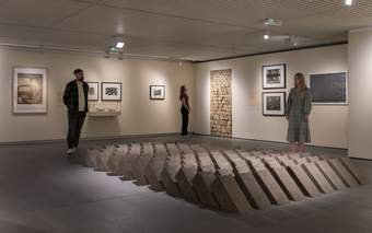 Three visitors examining various artworks in a Tate Liverpool's display. The central feature is a large three-dimensional geometric sculpture placed on the floor. The walls have framed artworks and photographs.