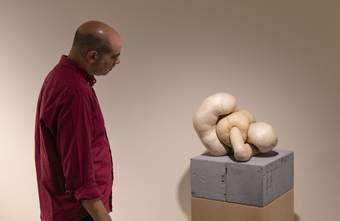 A person is closely examining a modern sculpture consisting of intertwining forms displayed on a pedestal made of breeze blocks.