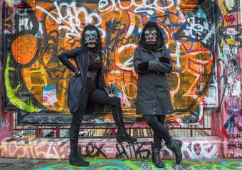 Two people wearing gorilla marks against a wall covered in graffiti.