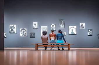 Photograph of 3 young people sitting on a bench looking up at a series of photographs on the Tate gallery wall.