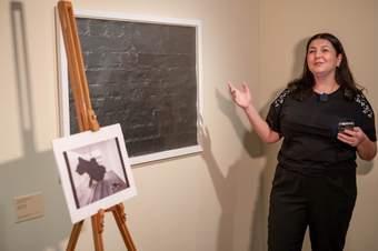 A photograph of a person talking in front of an artwork whilst holding out their hand, indicating towards the art on display on the left and an artwork on an easel..