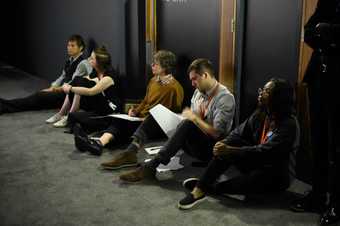 Five people sit on the floor at the edge of a dark room, their backs against a wall, appearing to watch something