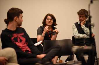 Two seated people turn to look at a third who is speaking and gesturing with their hands