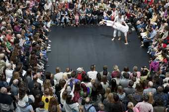 Two male dancers wearing white dance in the Turbine Hall with a crowd watching them forming a square. One is lifting the other