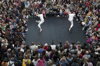 Two male dancers wearing white leap in the air in unison whilst being watched by a crowd