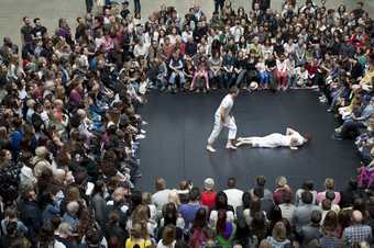 Two male dancers wearing white dance in the Turbine Hall. One is lying down, the other peering over him.