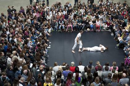 Two male dancers wearing white dance in the Turbine Hall. One is lying down, the other peering over him.