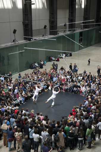 Two male dancers wearing white dance in the Turbine Hall with a crowd watching them forming a square