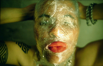 An individual whose head is wrapped in clear cling film with a hole for red-lipsticked lips