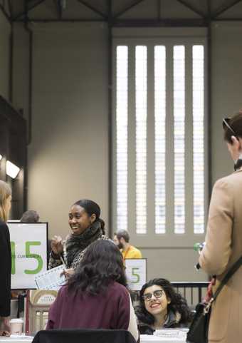 Young people smiling as they engage with visitors at the Alternative Careers Fair with the distinctive vertical windows of the Tate Modern Turbine Hall behind