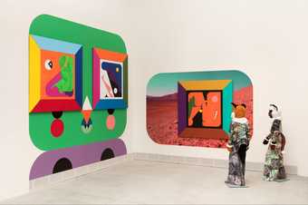 two standing sculptures with fox heads stand in a gallery space looking at two brightly coloured, geometric paintings