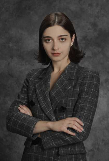 portrain photograph of a woman with glossy brown hair tucked behind her ears. arms are folded over each other and she is wearing a checkered blazer.
