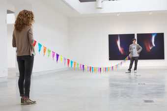 Two people stand in a gallery at a distance connected by a string of bunting