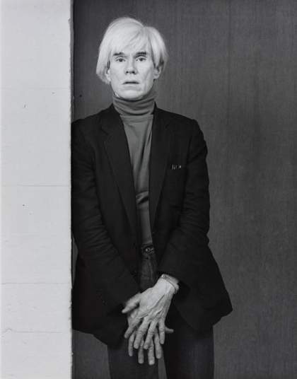 black and white portrain photograph of Andy warhol leaning against a wall. He is wearing a turtleneck and a blazer and his hands are over one another.