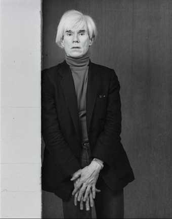black and white portrain photograph of Andy warhol leaning against a wall. He is wearing a turtleneck and a blazer and his hands are over one another.