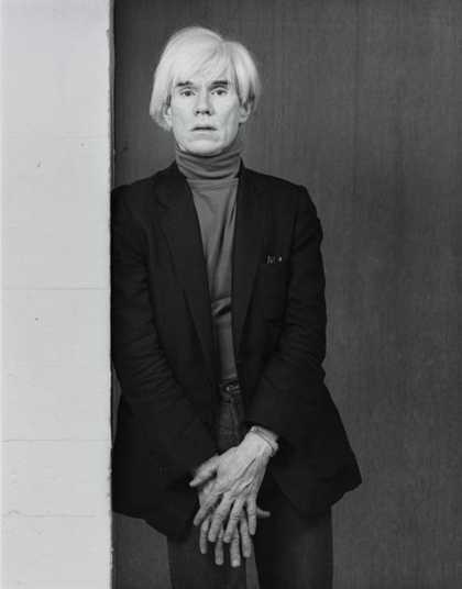 black and white portrain photograph of Andy warhol leaning against a wall. He is wearing a turtleneck and a blazer and his hands are over one another.