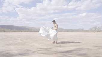 A person holds a white sheet around themselves which blows in the wind. They are in a desert with blue skies and small mountains in the distance.