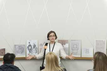 An archive explorer leads a discussion in the archive gallery with paper drawings stood up on a shelf behind her