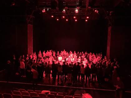 people in a theatre in a circle, in a black room lit by pink lighting