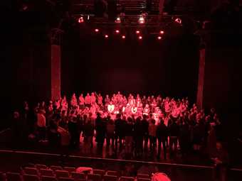 people in a theatre in a circle, in a black room lit by pink lighting 