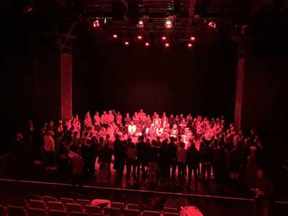 people in a theatre in a circle, in a black room lit by pink lighting